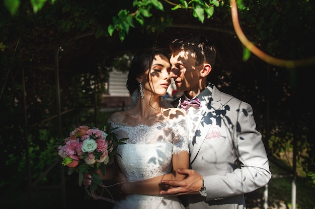 A loving couple of newlyweds gently hug in the shade of trees