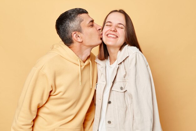 Loving couple woman and man wearing casual style clothing standing isolated over beige background expressing love husband kissing his wife