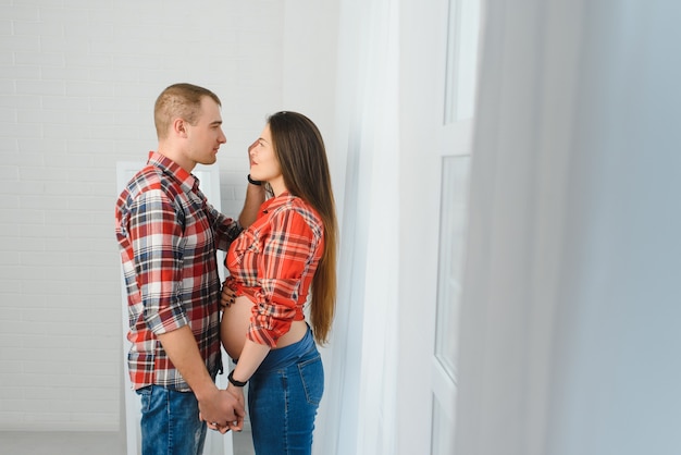 Loving man hugging his pregnant wife from behind standing near window at home, copy space