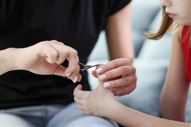 Loving mom cuts off tiny nails of little daughter at home sitting on sofa woman helps girl to