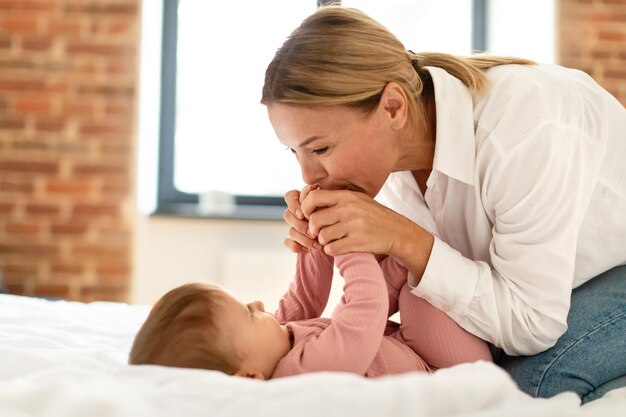 Loving mommy holding her baby daughter hands and kissing waking up her infant guirl after daytime sleep lying in bed