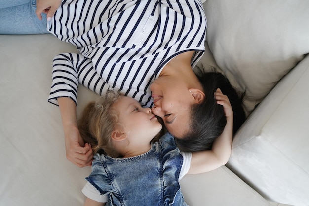 Loving mother lying on couch touching nose face with little daughter at home. Top view mom and kid girl relaxing on sofa, enjoy cute moment together. Happy motherhood, family bond, unconditional love.