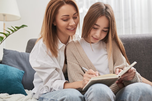 Loving mother writing in copybook and explaining homework to preteen daughter while sitting together on sofa during studies at home