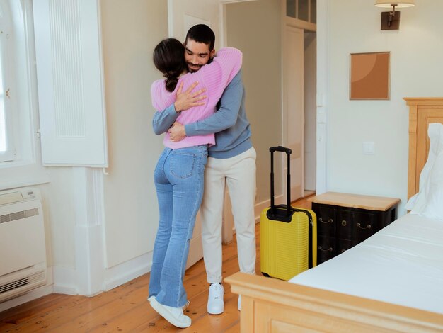 Loving spouses hugging standing with suitcase at hotel suite interior