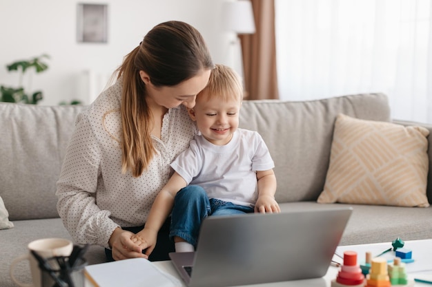 Loving young mom playing with son while working on laptop sitting on sofa and tickling child