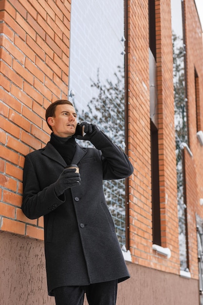 Photo low angle of adult businessman with paper cup of coffee to go making phone call near office building