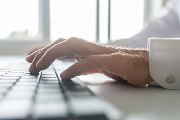 Low angle closeup view of businessman typing on computer keyboard with bright office windows in background.