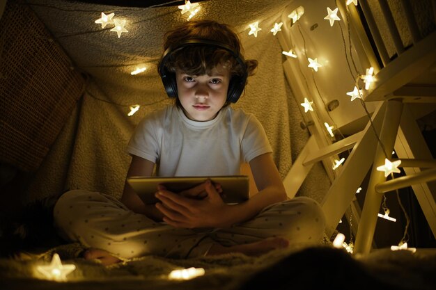 Low angle full body of boy in pajama and wireless headphones watching video on tablet sitting in playing tent decorated with glowing garland