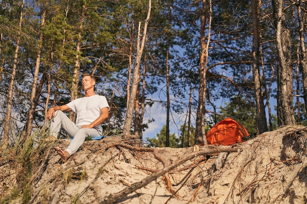 Low angle of happy mature sporty male sitting on cliff among nature while hiking with tent