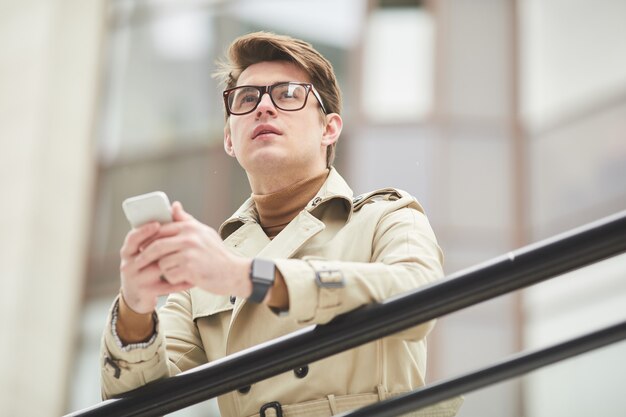 Low angle portrait of modern young businessman holding smartphone outdoors while leaning on railing and looking away in urban city setting, copy space