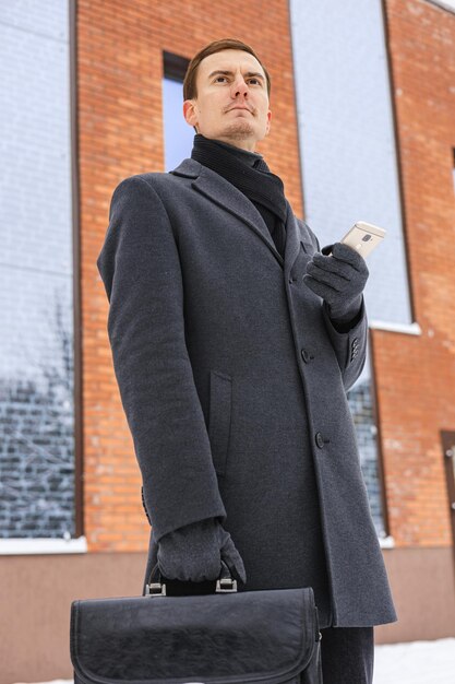 Photo low angle of serious businessman in winter coat browsing mobile phone while standing in city center