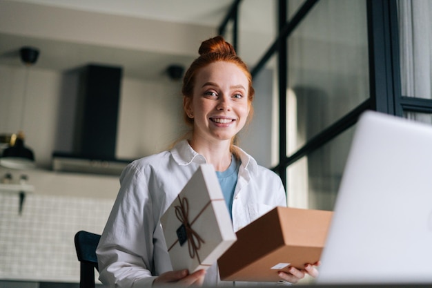 Low-angle shot of cheerful redhead young woman opening gift box with present during video call on laptop sitting at desk. Concept of leisure activity red-haired female at home during self-isolation.