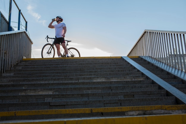 Low angle shot of a male muscular cyclist drinking water, standing near his bicycle on asummer day, copy space