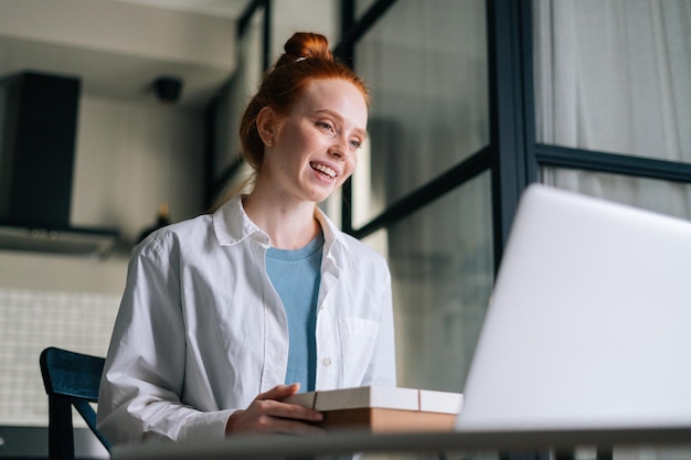 Low-angle view of cheerful redhead young woman opening gift box during video call via laptop webcam. Concept of leisure activity red-haired female at home.