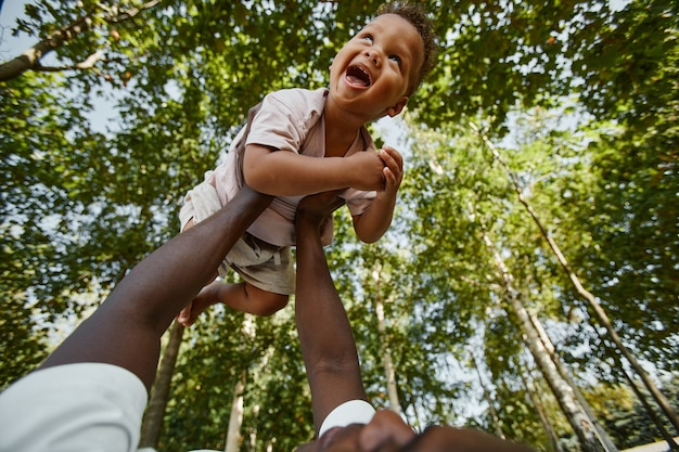 Low angle view at cute baby boy laughing happily while playing with father in park against trees cop...