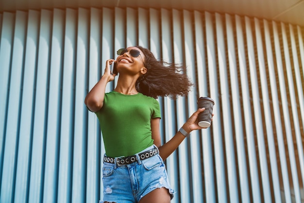 Low angle view of happy woman standing near wall with coffee while talking on mobile phone