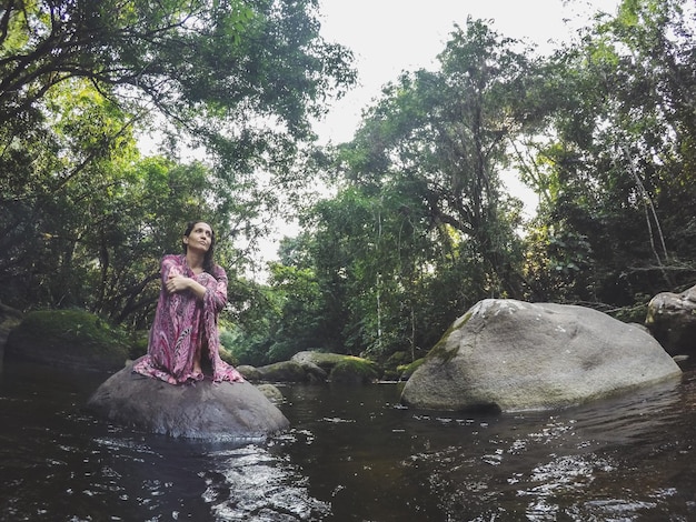 Photo low angle view of mid adult woman sitting on rock in lake at forest