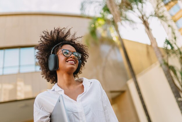 Low angle view of smiling Latin businesswoman holding laptop and eyeglasses near blurred building