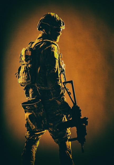 Low key studio shoot of army special forces soldier, commando fighter in mask, ballistic glasses, tactical helmet and battle uniform, holding short barrel service rifle, looking back over shoulder