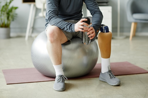 Low section closeup of man with prosthetic leg sitting on fitness ball and holding water bottle afte