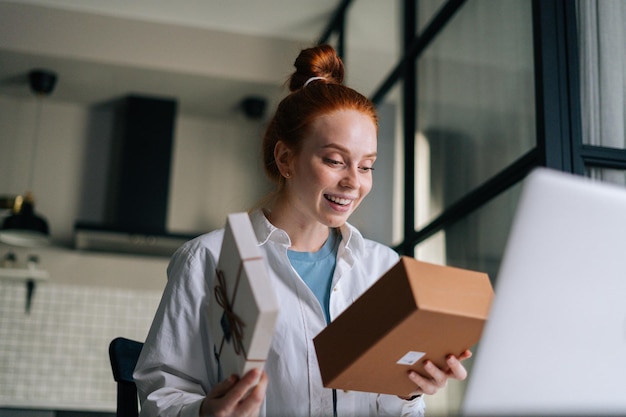 Lowangle shot of cheerful redhead young woman opening gift box with present during video call