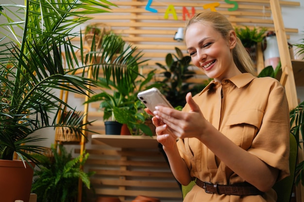 Lowangle view of cheerful female florist typing message to client and chatting with customer using mobile phone looking at smartphone screen