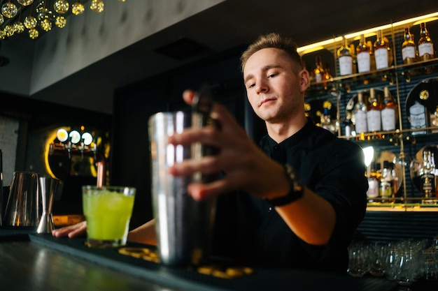 Lowangle view of confident bartender male making colorful alcoholic cocktail standing behind bar counter in modern dark nightclub