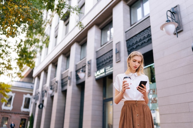 Lowangle view of fashionable beautiful female teenager chatting in smartphone looking to device screen holding cup of coffee outdoors