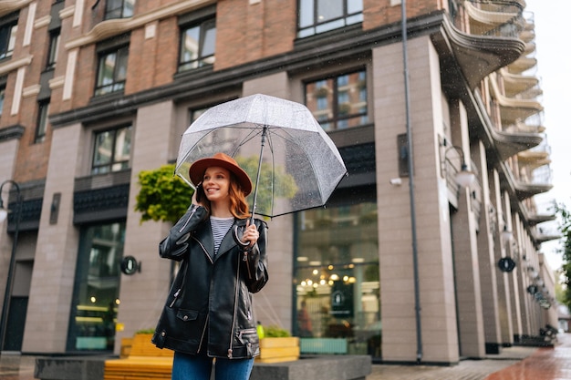 Lowangle view of happy young woman wearing elegant hat standing with transparent umbrella talking on smartphone in rain on European city street looking away