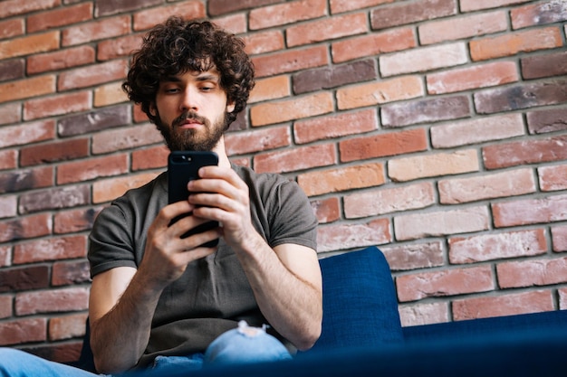 Lowangle view of serious bearded young man looking at smartphone screen browsing wireless internet on device sitting on sofa at home