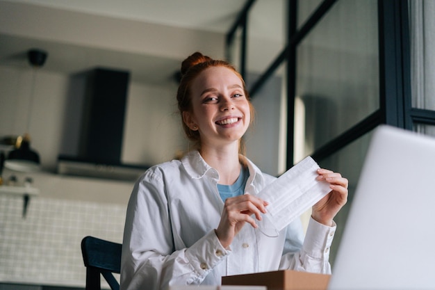 Lowangle view of surprised cheerful redhead young woman opening gift box and takes out medical mask during video call via laptop webcam Concept of leisure activity redhaired female at home