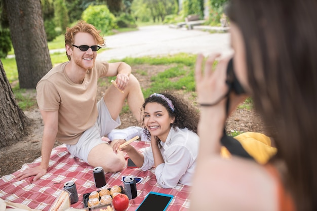 Lucky moment. Young girl taking photos of smiling relaxed friends having rest on picnic on warm day