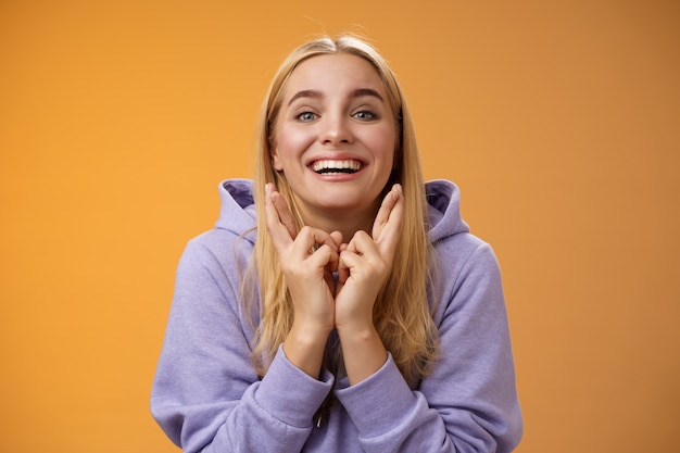 Lucky optimistic cute positive smiling blond girl grinning excited cross fingers making wish dreaming desire come true anticipating good news praying orange background waiting results.