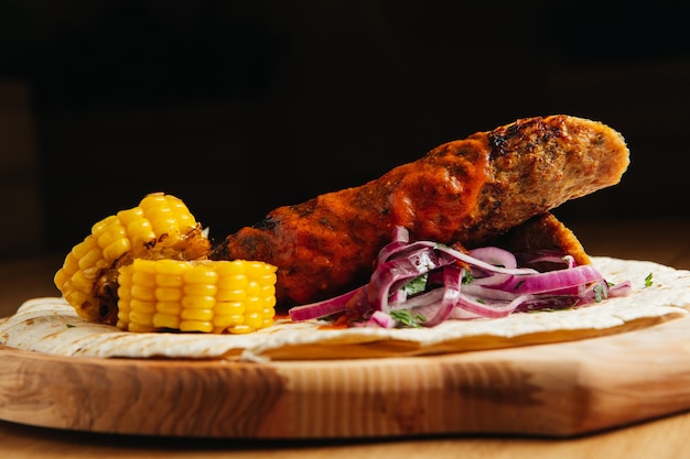 Lula kebab with pita bread, sauce, grilled corn and onion on a wooden board