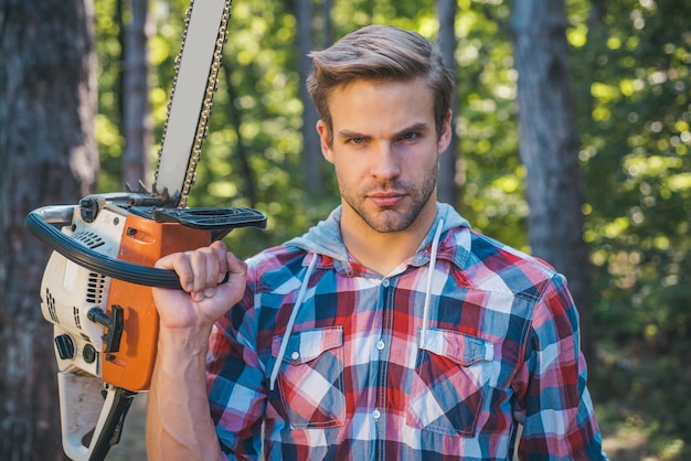 Lumberjack worker standing in the forest with chainsaw Chainsaw Stylish young man posing like lumberjack Woodcutter with chainsaw on sawmill Harvest of timber