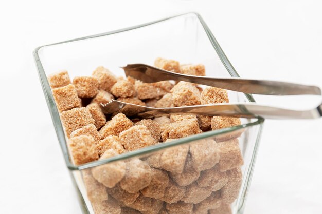 Lumps of brown sugar and tongs in a clear glass jar. Close-up. White background.