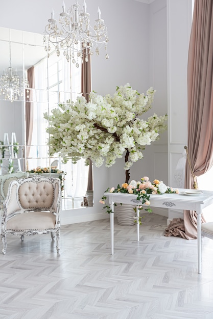 Luxurious bright spacious guest room with beautiful chic furniture a huge floor-to-ceiling window in a royal style is decorated with green plants, white walls with stucco and a fireplace