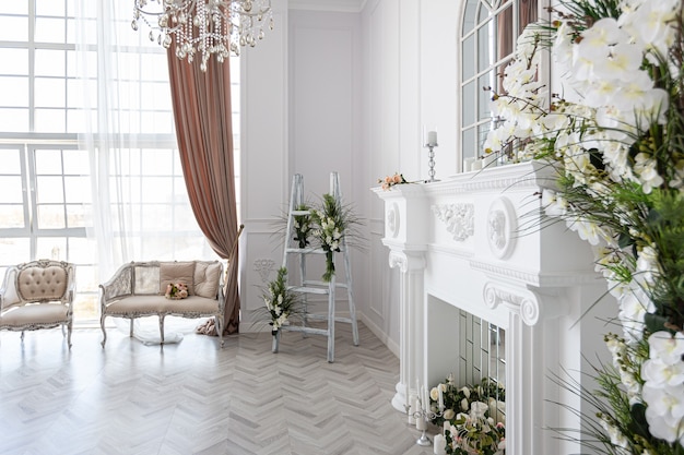 Luxurious bright spacious guest room with beautiful chic furniture a huge floor-to-ceiling window in a royal style is decorated with green plants, white walls with stucco and a fireplace