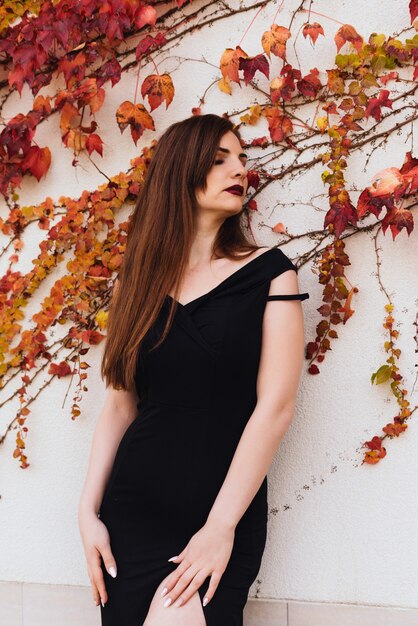 Luxurious long-haired young woman in black dress posing against wall in the open air, enjoying rest