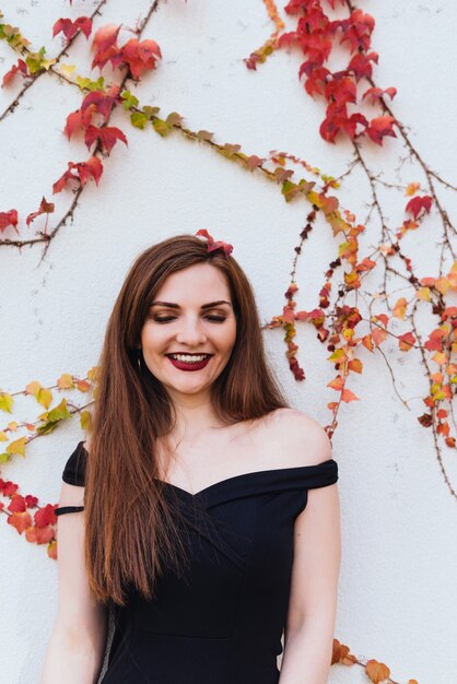 Luxurious long-haired young woman in black dress posing against wall outdoors and smiling