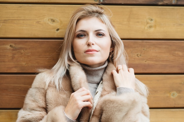 Luxury blond woman with blue eyes and long hair in fur coat 30 years old on wood wall.