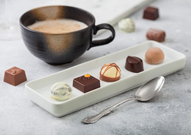 Luxury Chocolate candies in white porcelain plate with cup of cappuccino coffee and silver spoon on light background