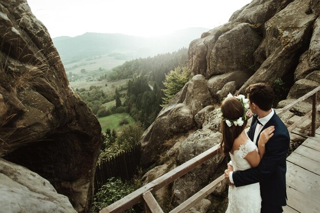 Photo luxury happy bride and stylish groom holding each other and looking on background of sunny rocks in amazing mountains