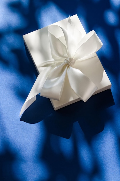 Luxury holiday white gift box with silk ribbon and bow on blue background luxe wedding or birthday present