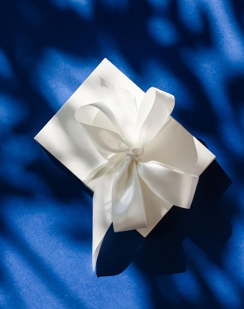 Luxury holiday white gift box with silk ribbon and bow on blue background luxe wedding or birthday present