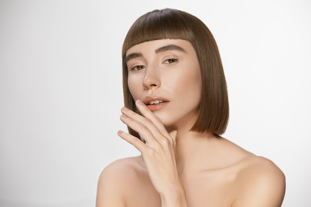 Luxury woman with fresh clean skin and stylish haircut with bangs, sensual pretty woman touching her glossy lips having nude daily make-up and shiny straigh hair