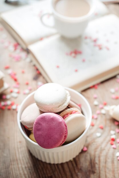 Macaroons in a bowl and a cup of coffee a book on the background of small hearts on a wooden background top view