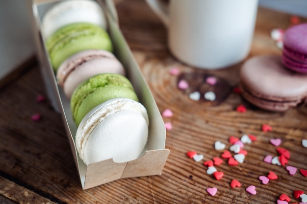 Macaroons in a box a cup of coffee a milk jug against the background of small hearts on a wooden background top view