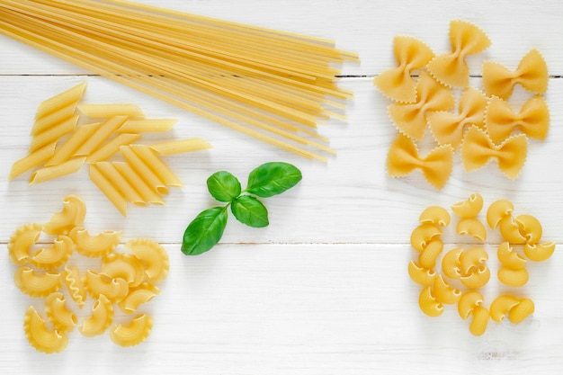 Maccheroni raw spaghetti farfalle pipe rigatoni penne rigate cresta di gallo leaf of basil on white wooden board background top view space to copy textxD