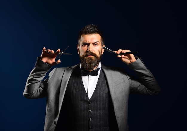 Macho in formal suit cuts and shaves beard and moustache. Business reduction concept. Businessman with serious face on dark blue background. Man with long beard holds shaving razor and scissors.